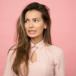 A young woman with a playful uncertain expression against a pink background.