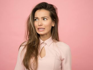 A young woman with a playful uncertain expression against a pink background.